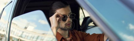 young stylish man with trendy accessories posing behind steering wheel, touching sunglasses, banner