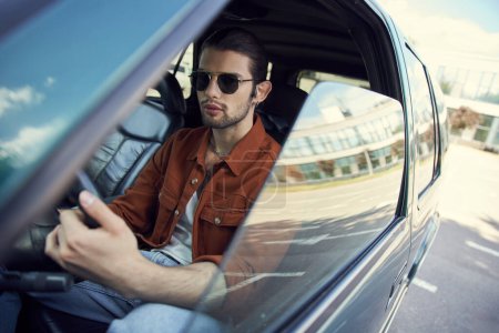 beau jeune modèle masculin en chemise marron élégant avec des lunettes de soleil au volant, chauffeur sexy
