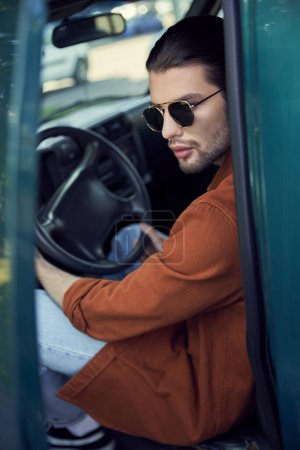 llamativo hombre elegante con gafas de sol en camisa marrón en su coche con las manos en el volante