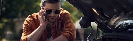 Foto de Hombre guapo con gafas llamando a su aseguradora y mirando la campana del motor abierto de su coche, pancarta - Imagen libre de derechos