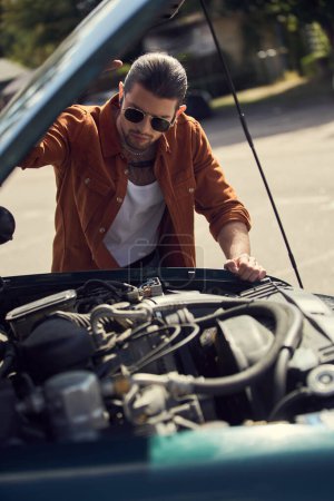attraktiver junger Mann in braunem Hemd mit Accessoires, der aufmerksam auf den Motor seines Autos blickt