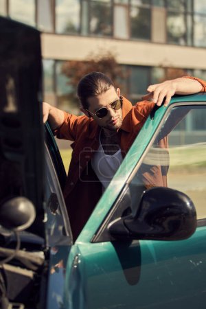 vertikale Aufnahme eines attraktiven sexy männlichen Modells mit stylischer Sonnenbrille, das neben seinem Auto posiert, Mode