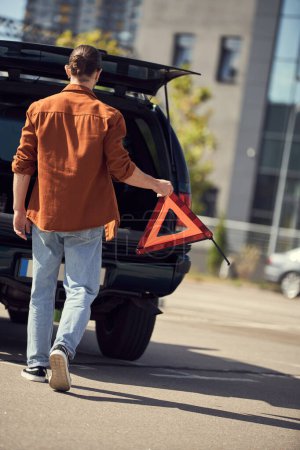 Téléchargez les photos : Vue arrière du jeune homme en chemise marron marchant vers sa voiture avec triangle d'avertissement dans les mains, chauffeur sexy - en image libre de droit