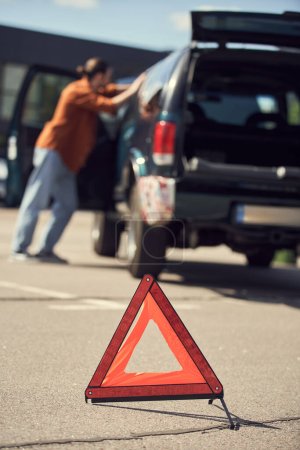 Téléchargez les photos : Photo floue du jeune homme en tenue urbaine stylée debout à côté de sa voiture, triangle d'avertissement - en image libre de droit