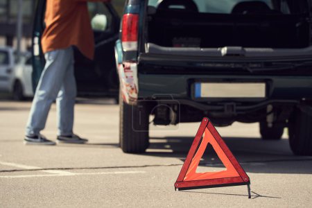 vista recortada de un hombre joven y elegante de pie junto a su coche con triángulo de advertencia, foto borrosa
