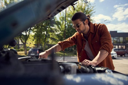 junger Mann mit Sonnenbrille und Bart in braunem, stylischem Hemd und Jeans checkt seinen Automotor