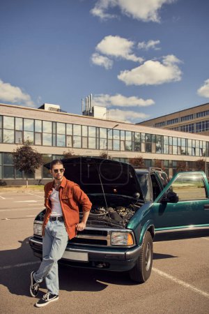 vertical shot of appealing stylish man in brown shirt with sunglasses posing next to his car