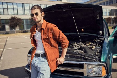 eye catching sexy driver in stylish brown shirt posing next to his car with opened engine hood