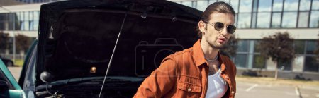 hombre sexy de buen aspecto en camisa marrón elegante posando junto a su coche con campana del motor abierto, pancarta