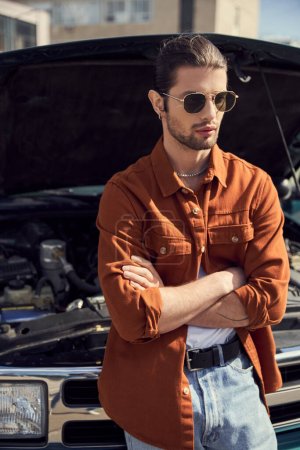stattliche auffällige männliche Modell in stilvoller Kleidung posiert neben seinem Auto mit gekreuzten Armen auf der Brust
