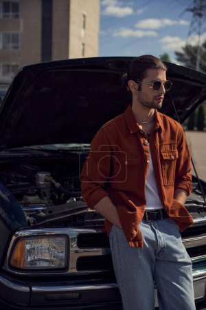 tiro vertical de joven guapo en camisa marrón con gafas de sol posando cerca de su coche roto