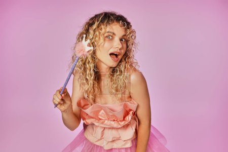 curly haired blonde woman in pink attire of tooth fairy holding magic wand and looking at camera