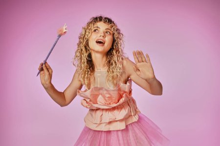 blonde curly haired woman with magic wand posing in pink tooth fairy costume on pink backdrop Poster 676831580