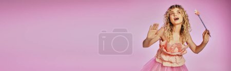 Foto de Hermosa mujer de pelo rizado con las manos levantadas y varita mágica posando sobre fondo rosa, bandera - Imagen libre de derechos
