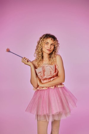 joyous pretty woman in pink outfit of tooth fairy holding magic wand with slightly crossed arms