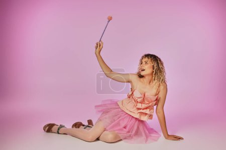 alegre rubia con pelo rizado posando sobre fondo rosa en traje de hada de los dientes con varita mágica