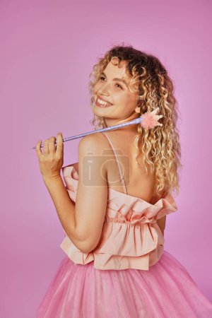 jolly beautiful woman in pink costume of tooth fairy holding magic wand and looking away happily