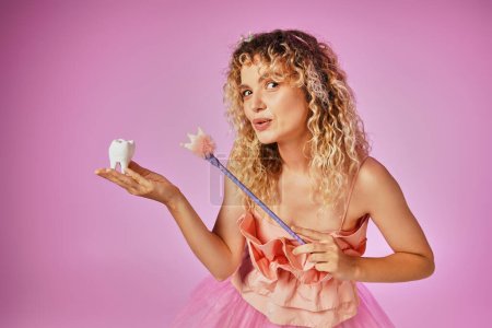 lovely blonde woman in tooth fairy costume playfully casting spell on baby tooth with her magic wand