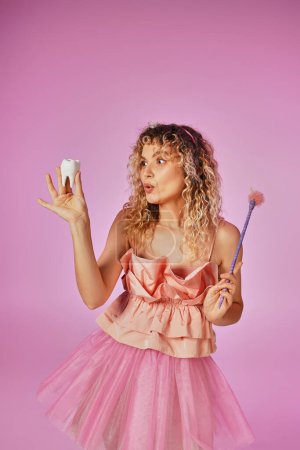 Photo for Amazed curly haired woman in tooth fairy attire with headband and magic wand looking at baby tooth - Royalty Free Image