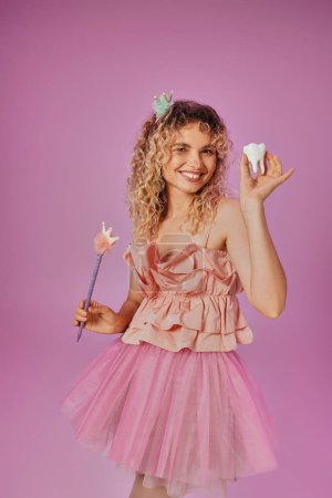cheerful lovely woman in tooth fairy costume holding magic wand and baby tooth and smiling at camera