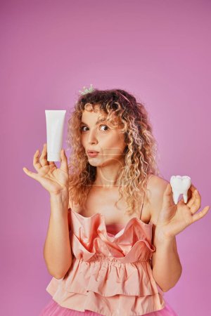 Photo for Amazed blonde woman in pink costume holding baby tooth and tooth paste and looking at camera - Royalty Free Image