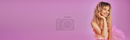 Photo for Joyous lovely woman in pink costume smiling sincerely at camera with hand up to face, banner - Royalty Free Image
