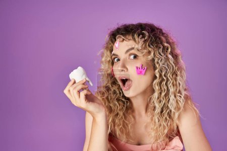 portrait of amazed blonde woman with face stickers holding baby tooth and looking shocked at camera