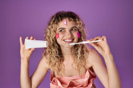 Téléchargez les photos : Fée des dents blonde souriante en costume rose avec autocollants visage tenant brosse à dents et pâte dentaire - en image libre de droit
