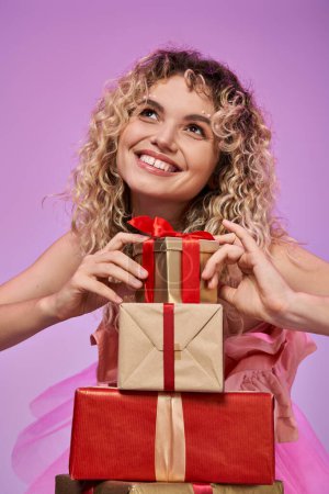 portrait of dreamy pretty woman with curly hair touching pile of gifts with fingers and looking away