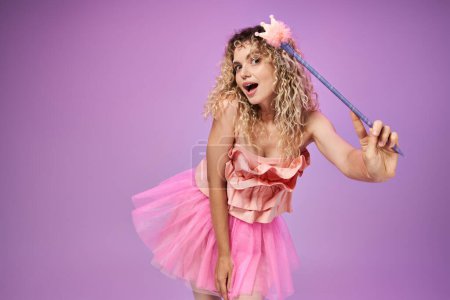 amazed woman in pink tooth fairy costume with magic wand in hand casting spell and looking at camera
