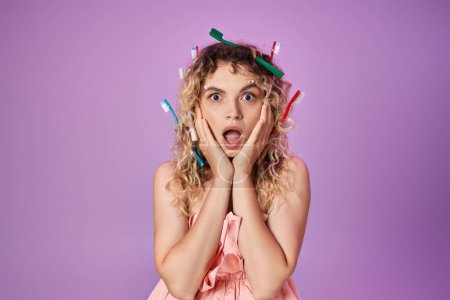 Schockierte Frau in rosa Kleid und Zahnbürsten im Haar, die Hände zu Wangen erhoben, Zahnfee-Konzept