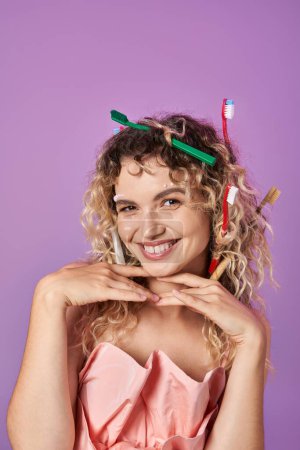 Téléchargez les photos : Joyeuse femme en robe rose et brosses à dents dans ses cheveux, les mains sous le menton, concept de fée des dents - en image libre de droit
