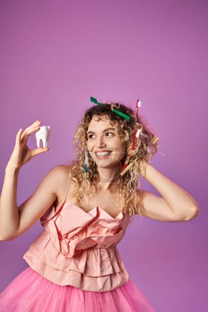 cheerful woman in pink dress with toothbrushes in her hair posing with tooth on pink backdrop