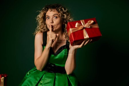 pretty elf posing in green dress with present in hands on green backdrop showing silence gesture