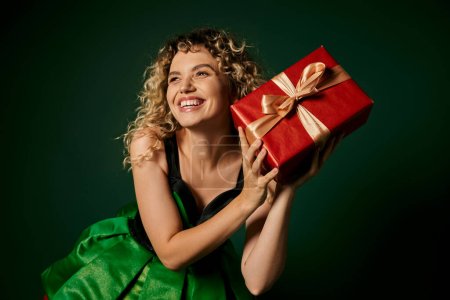 fröhliche Elfe in grünem Kleid posiert mit Geschenk in der Hand vor dunkelgrünem Hintergrund und lächelt glücklich