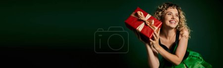 happy new year elf in green dress posing with present in her hands on dark green backdrop, banner
