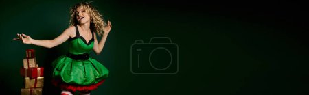 Téléchargez les photos : Femme joyeuse en costume d'elfe geste avec les bras posant près pile de cadeaux sur fond vert, bannière - en image libre de droit