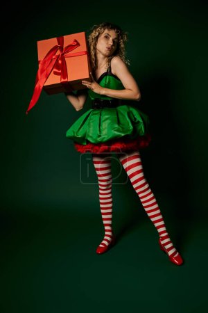 dreamy new year elf with pouted lips in green dress and striped stockings holding huge gift