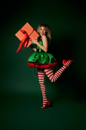 Téléchargez les photos : Heureux nouvel elfe bouclé année debout sur une jambe et tenant énorme présent dans les mains souriant à la caméra - en image libre de droit