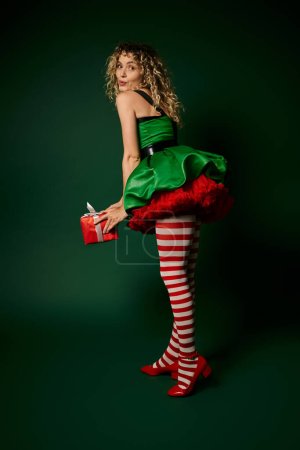 jolly woman in new year elf costume posing with pouted lips and holding huge present in hands