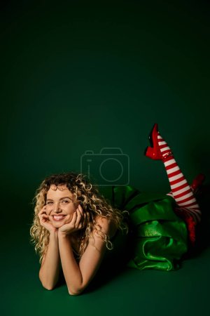 glückliche Frau, die als Neujahrselfe gekleidet auf dem Boden posiert, die Beine leicht erhöht auf grünem Hintergrund