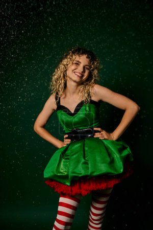 cheerful woman in green festive dress smiling and holding hands akimbo, new year elf concept