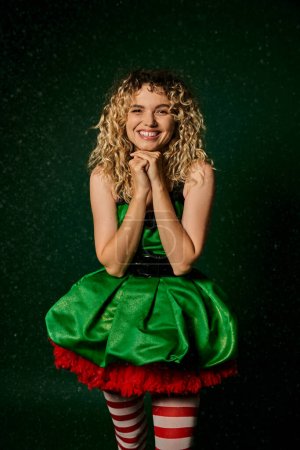 Photo for Smiling new year elf in festive dress and stockings looking cheerfully at camera, hands to face - Royalty Free Image