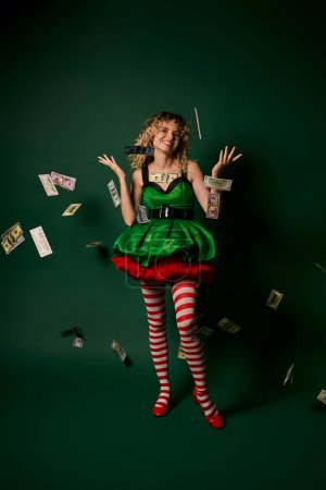 alegre rizado mujer vestida como nuevo año elfo de pie bajo el dinero lluvia alegremente mirando a la cámara