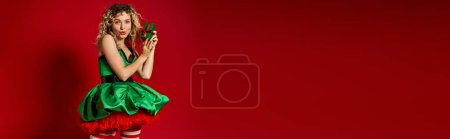 Photo for Astonished new year elf in green dress with pouted lips holding gift and looking at camera, banner - Royalty Free Image