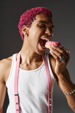 Porträt eines jungen Mannes mit rosafarbenen Hosenträgern, der rosafarbene, leckere Donuts isst, Mode und Stil