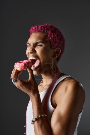 voguish gutaussehender Mann mit rosa Haaren und Accessoires essen köstlichen rosa Donut, Mode und Stil