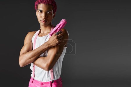 modisch gut aussehendes männliches Model mit rosa Haaren posiert mit Spielzeugpistole und schaut ernst in die Kamera