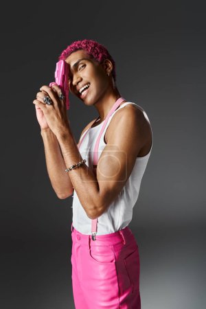 beau gai homme avec les cheveux bouclés rose posant avec pistolet jouet rose et souriant à la caméra