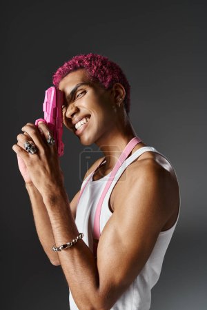 cheerful male model with curly pink hair posing in profile with pink toy gun and smiling at camera